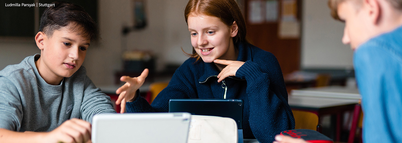 Drei Schüler sitzen im Klassenzimmer an einem Tisch, diskutieren und schauen auf ein Tablet